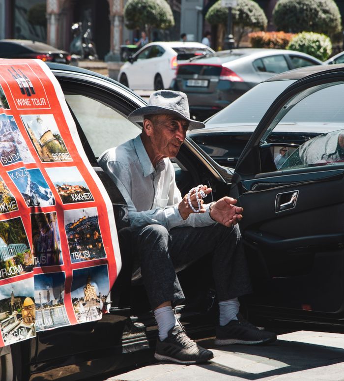 services-04 Senior man in hat sits in car displaying a travel poster. Urban street scene.
